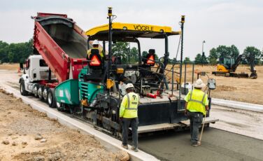 Two construction follow behind a concrete paving vehicle along a newly built stretch of road.